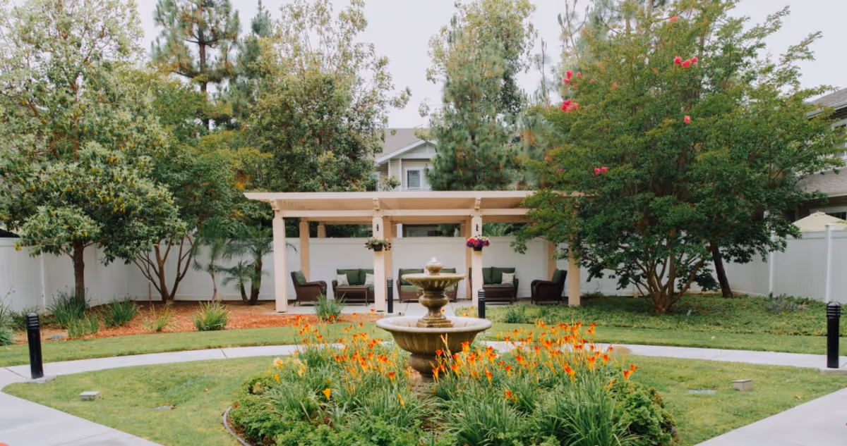 Outdoor courtyard featuring a central tiered fountain surrounded by flower beds, paved paths, a pergola with seating, and trees.