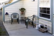 Outdoor patio area with two plastic chairs and a small table between them, placed on a concrete surface next to a light-colored building with a window. There is a plant on a stand and a small pet cage on a metal rack near the window.
