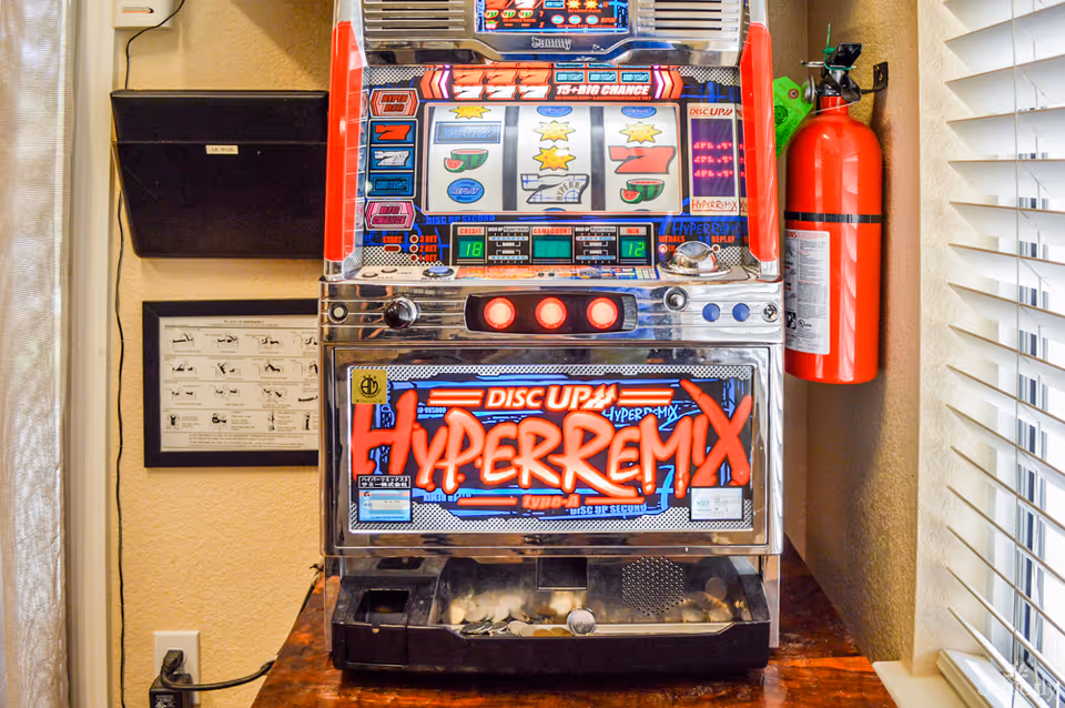 A slot machine labeled 'Hyper Remix' sits on a wooden surface next to a red fire extinguisher mounted on the wall. The machine has colorful lights and displays, with some coins visible in the tray at the bottom. There is a framed instructional poster on the wall behind the machine and a window with white blinds to the right.