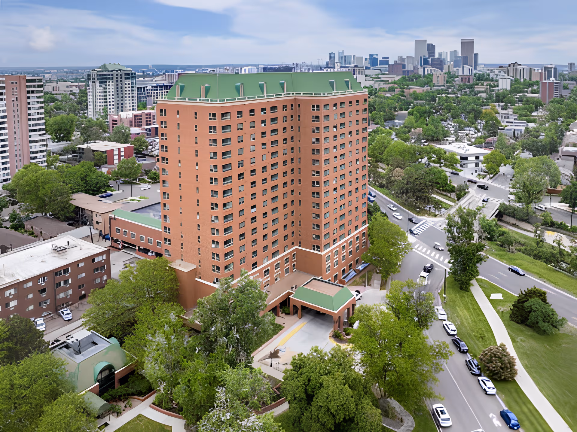 Aerial view of a tall brick building with a green roof surrounded by trees and roads, with a city skyline in the background under a partly cloudy sky.