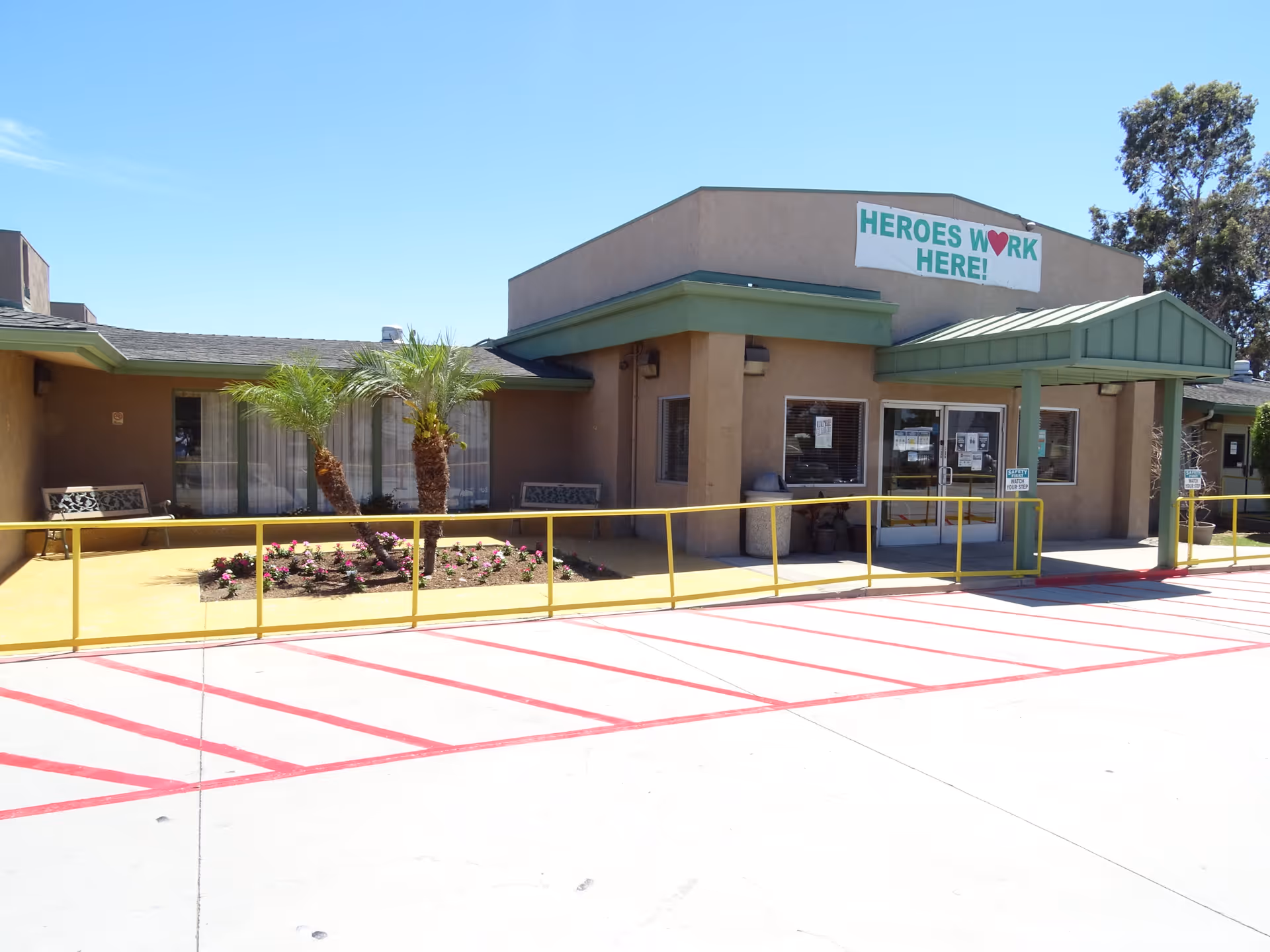 Exterior view of Rosecrans Care Center building entrance with a covered walkway, yellow railings, palm trees, flower bed, and benches under a clear blue sky. A banner above the entrance reads 'HEROES WORK HERE!'