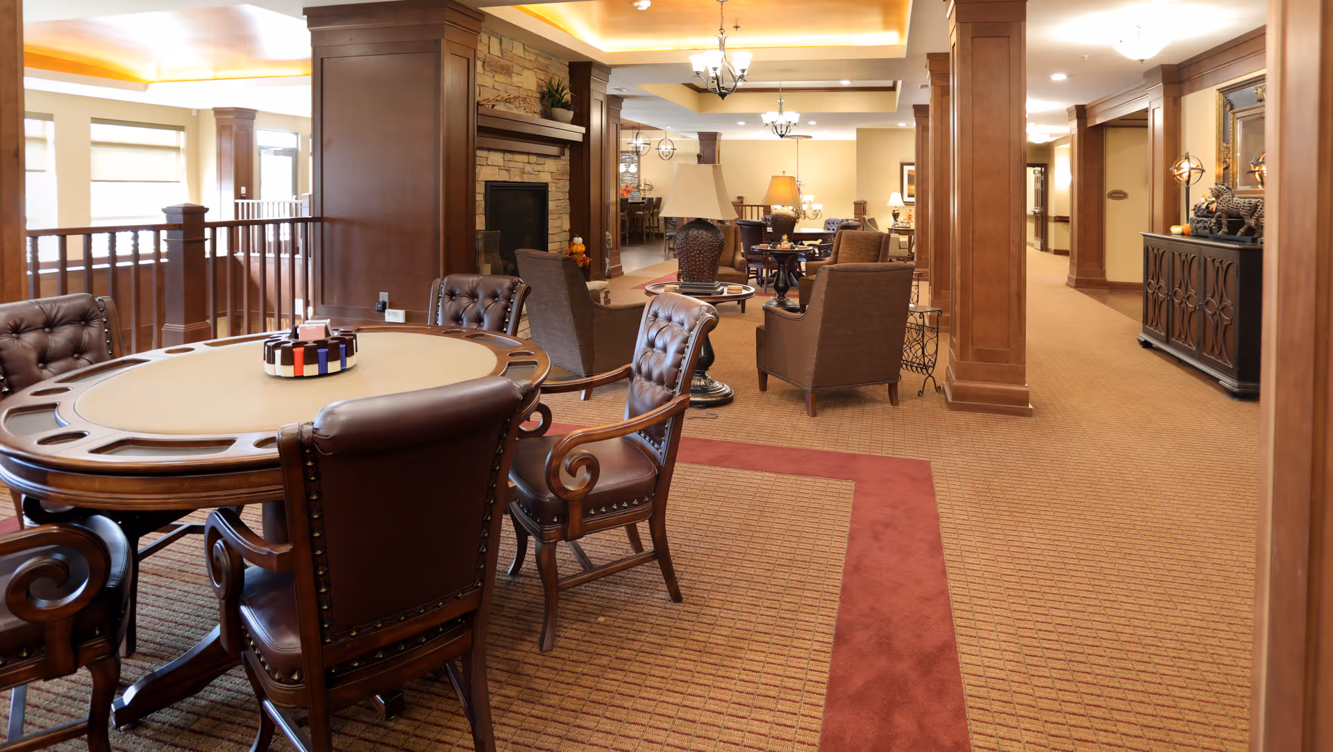 Warm, wood-paneled senior living common area with a card table and leather chairs, lounge seating, and a fireplace.