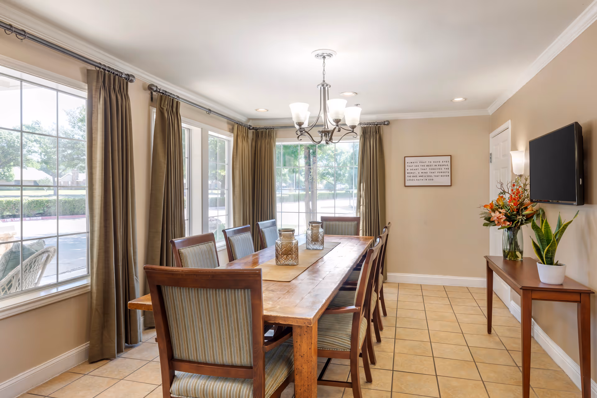 A bright dining room with a long wooden table surrounded by eight chairs with striped upholstery. Large windows with brown curtains let in natural light. A chandelier with five lights hangs above the table. On the right wall, there is a flat-screen TV mounted above a wooden console table decorated with a vase of flowers and a potted plant. A framed inspirational quote is also visible on the wall.