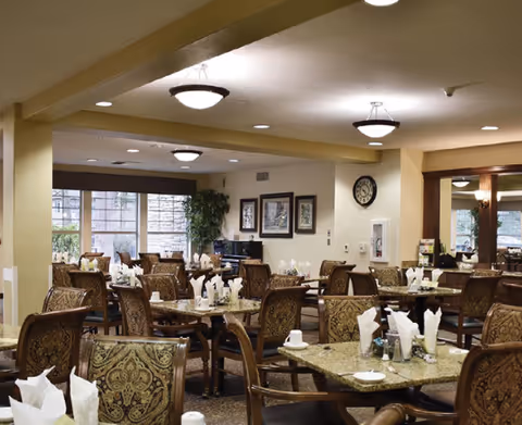 A dining room with multiple tables and chairs arranged neatly. Each table has white napkins, cups, and condiments. The room has warm lighting with ceiling fixtures, framed pictures on the wall, and large windows letting in natural light. There is a plant in the corner and a cozy, inviting atmosphere.
