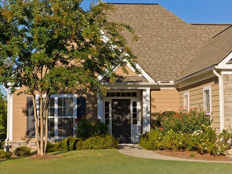 Exterior view of a single-story residential building with beige siding, a dark front door, white trim, a gabled roof, and surrounded by green bushes and a tree on a sunny day.