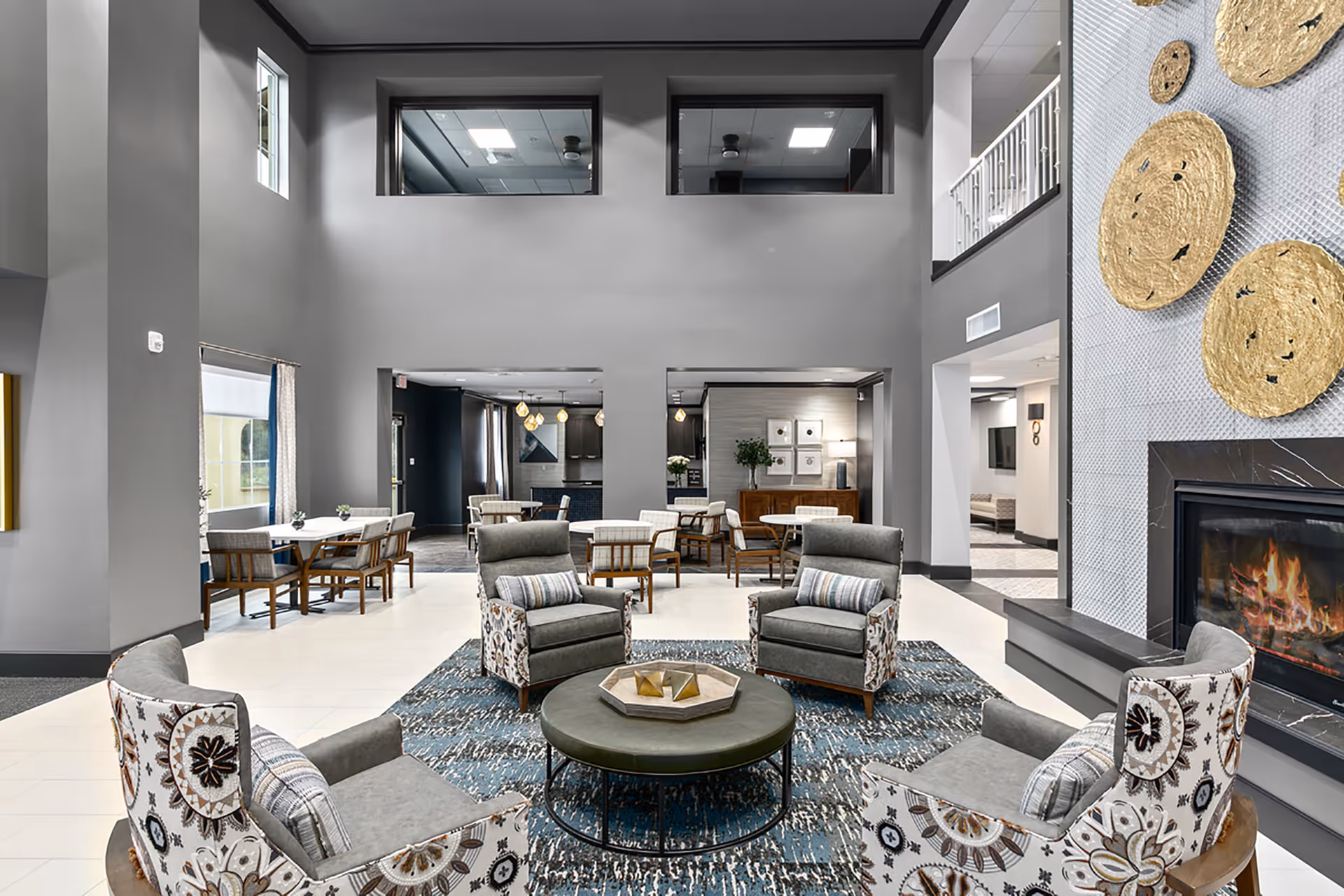 A spacious and modern living room area in Regency Fallbrook featuring four patterned armchairs arranged around a round green ottoman on a blue patterned rug. The room has high ceilings, large windows, and a fireplace with decorative golden circular wall art above it. In the background, there are dining tables and chairs, and a kitchen area with pendant lights.