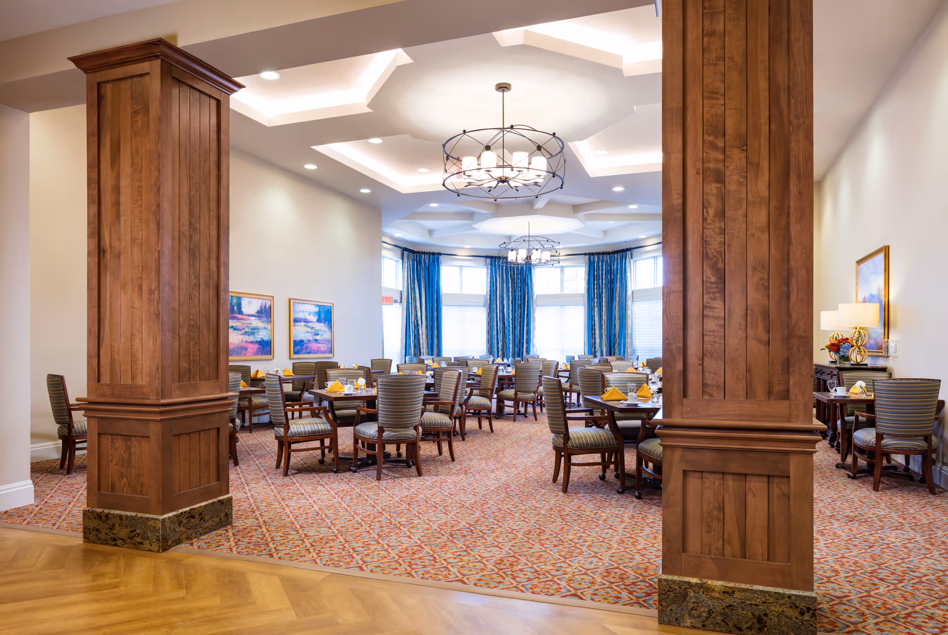 A spacious dining room with multiple round tables and chairs arranged neatly. Each table is set with yellow napkins and glassware. The room features large wooden columns, patterned carpet, and large windows with blue curtains allowing natural light to fill the space. Modern chandeliers hang from the ceiling, and framed artwork decorates the walls.