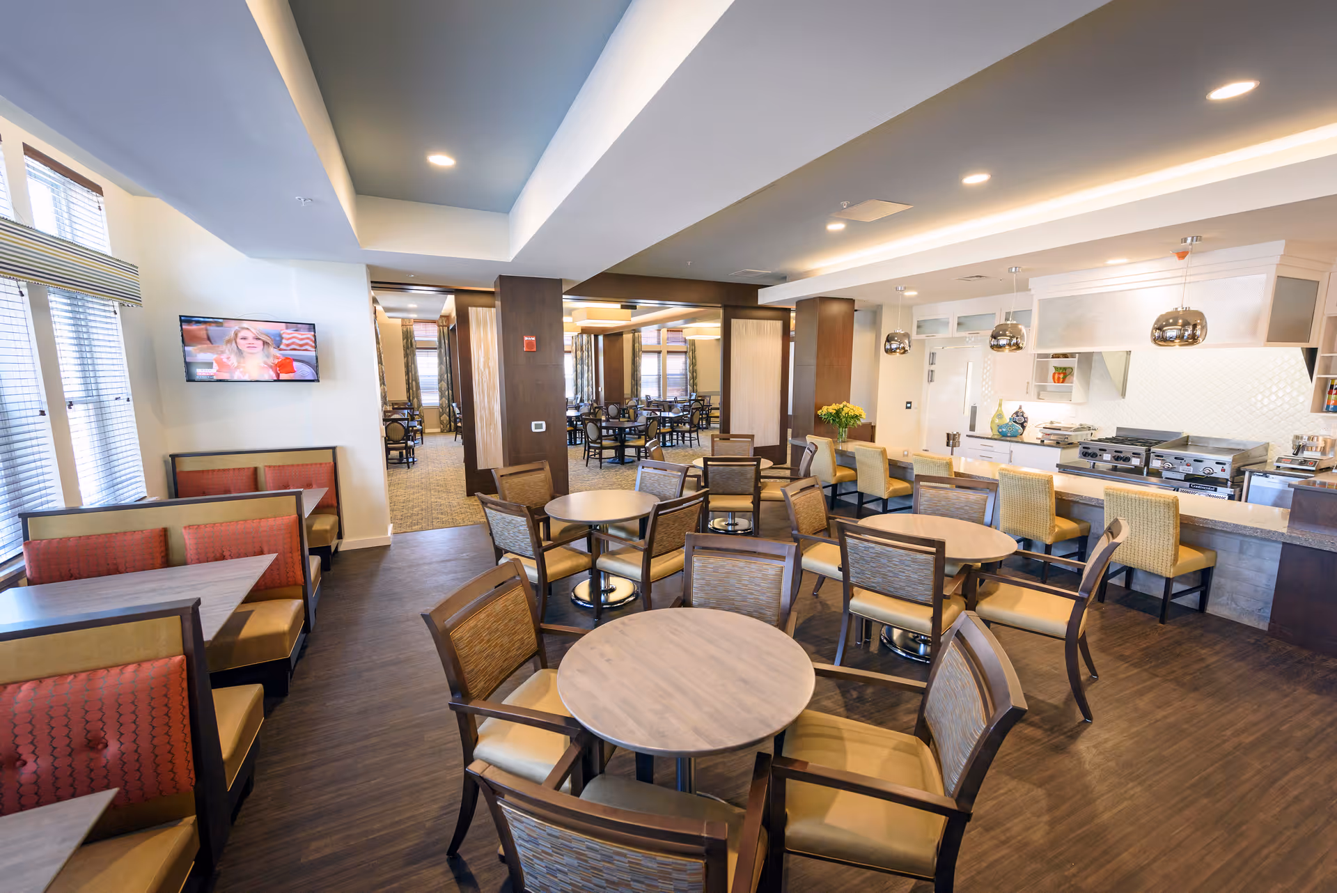 A bright and spacious dining area with multiple round tables and chairs arranged neatly. There are booth seating areas with red cushions along the left wall under windows with blinds. A flat-screen TV is mounted on the wall above the booths. The right side features a kitchen counter with bar stools and modern pendant lights hanging above. The room has wood flooring and recessed lighting in the ceiling.