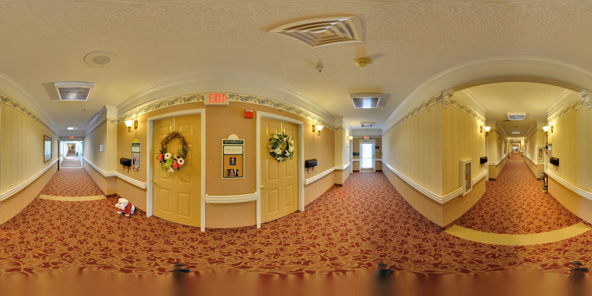 A long, carpeted hallway in a senior living facility with beige walls and floral-patterned red carpet. The hallway features multiple doors decorated with wreaths, wall-mounted lights, and exit signs. There are handrails along the walls and mail slots next to some doors. The hallway extends in both directions with natural light visible at the far ends.