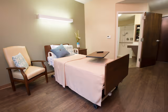 A healthcare facility room with a hospital bed covered in light pink bedding, a beige armchair with a patterned cushion, a bedside table with a vase of flowers, and an open door leading to a bathroom with a shower and sink.