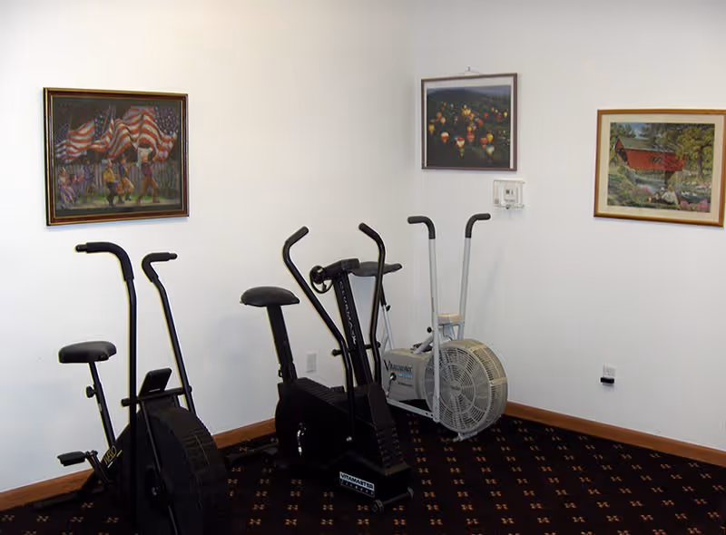 A small exercise room with three stationary exercise bikes placed against white walls. The floor is carpeted with a dark patterned carpet. Three framed paintings hang on the walls above the exercise equipment.