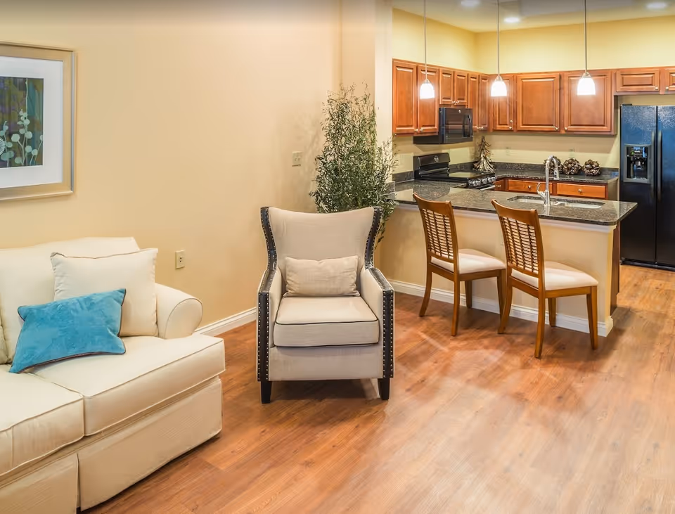 Open living area with a sofa, armchair, and a kitchen island with two bar stools under pendant lights.