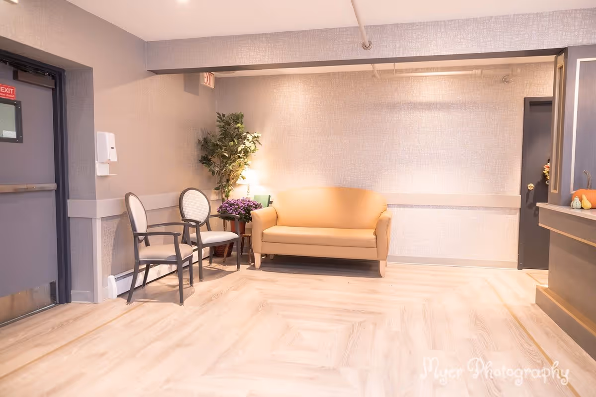 A waiting area with a beige loveseat, two white cushioned chairs with dark frames, a potted plant, a small table with a purple flower arrangement, and a lamp. The walls are light-colored with a textured pattern, and the floor has a light wood finish. There is an exit door on the left and a dark door on the right. A reception counter is partially visible on the right side of the image.