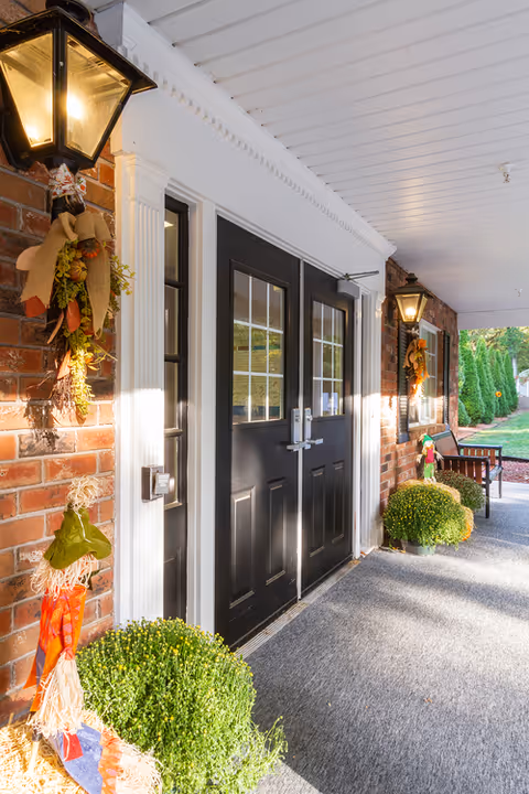 Covered entrance with double black doors, wall lanterns, fall decorations, potted mums and a bench along a brick facade.