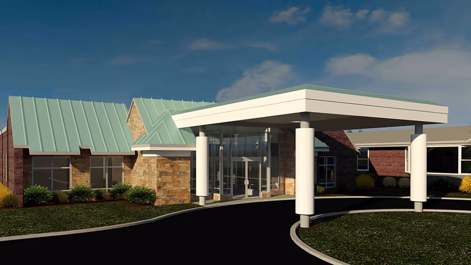 Front entrance of a single-story brick and stone assisted living building with a covered porte-cochère and green metal roofs.