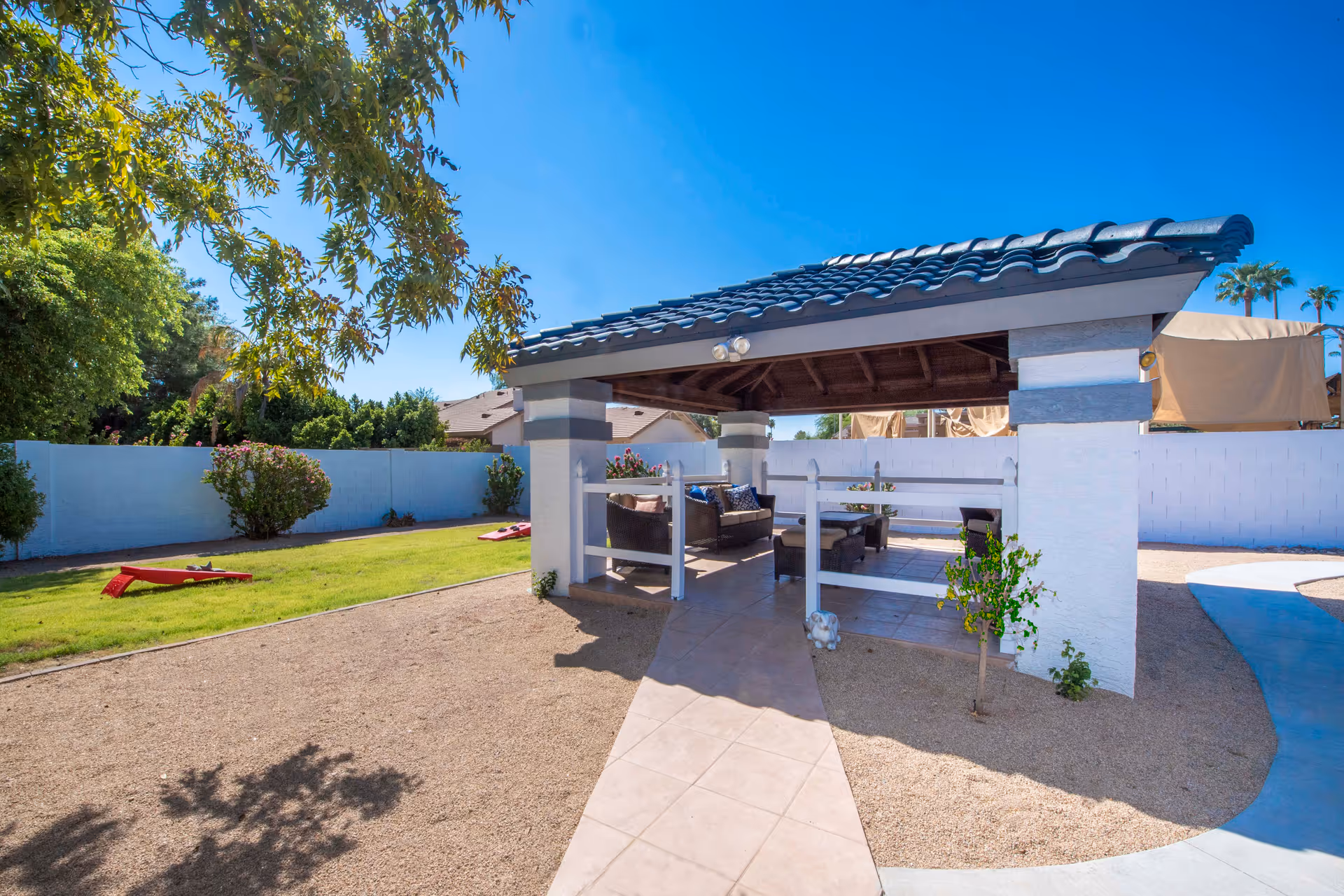 Sunlit outdoor courtyard with a covered patio pavilion, seating area, lawn and a tiled walkway.