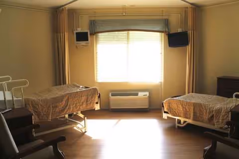 Two single beds in a shared senior living bedroom with a window and wall-mounted TVs.