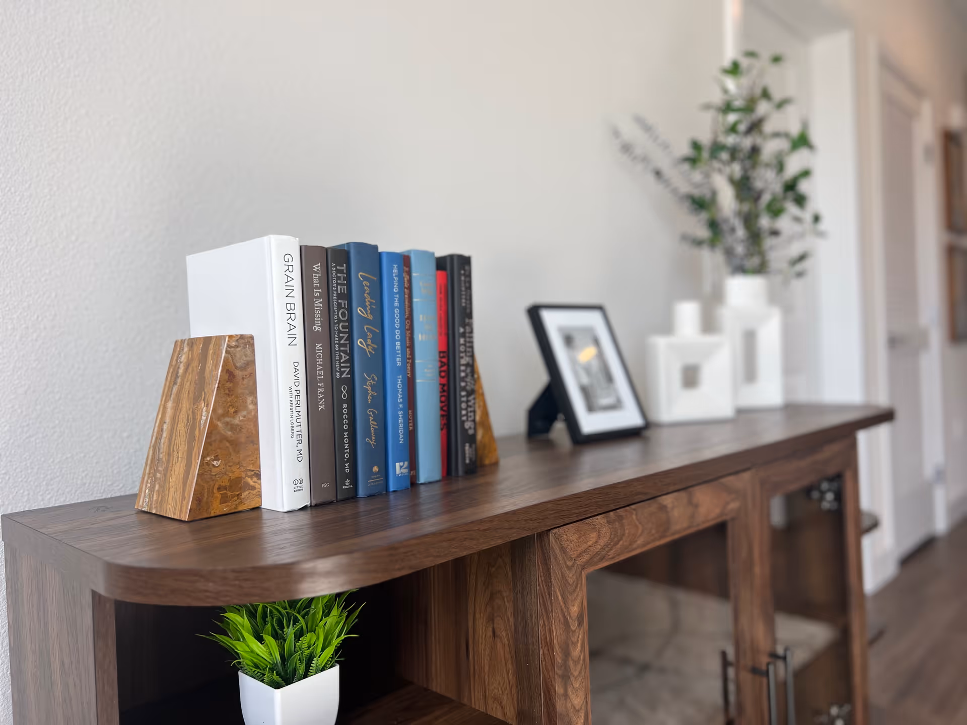 A wooden cabinet with a row of books held by stone bookends on top. Next to the books is a small framed photo and three white decorative vases with green foliage. Below the cabinet top is a small green potted plant. The background shows a white wall and a hallway with doors.