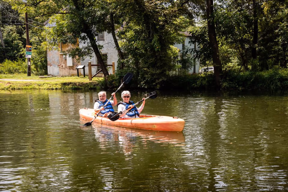 Two elderly people wearing life jackets paddling together in an orange kayak on a calm body of water surrounded by trees and greenery, with a building visible in the background.