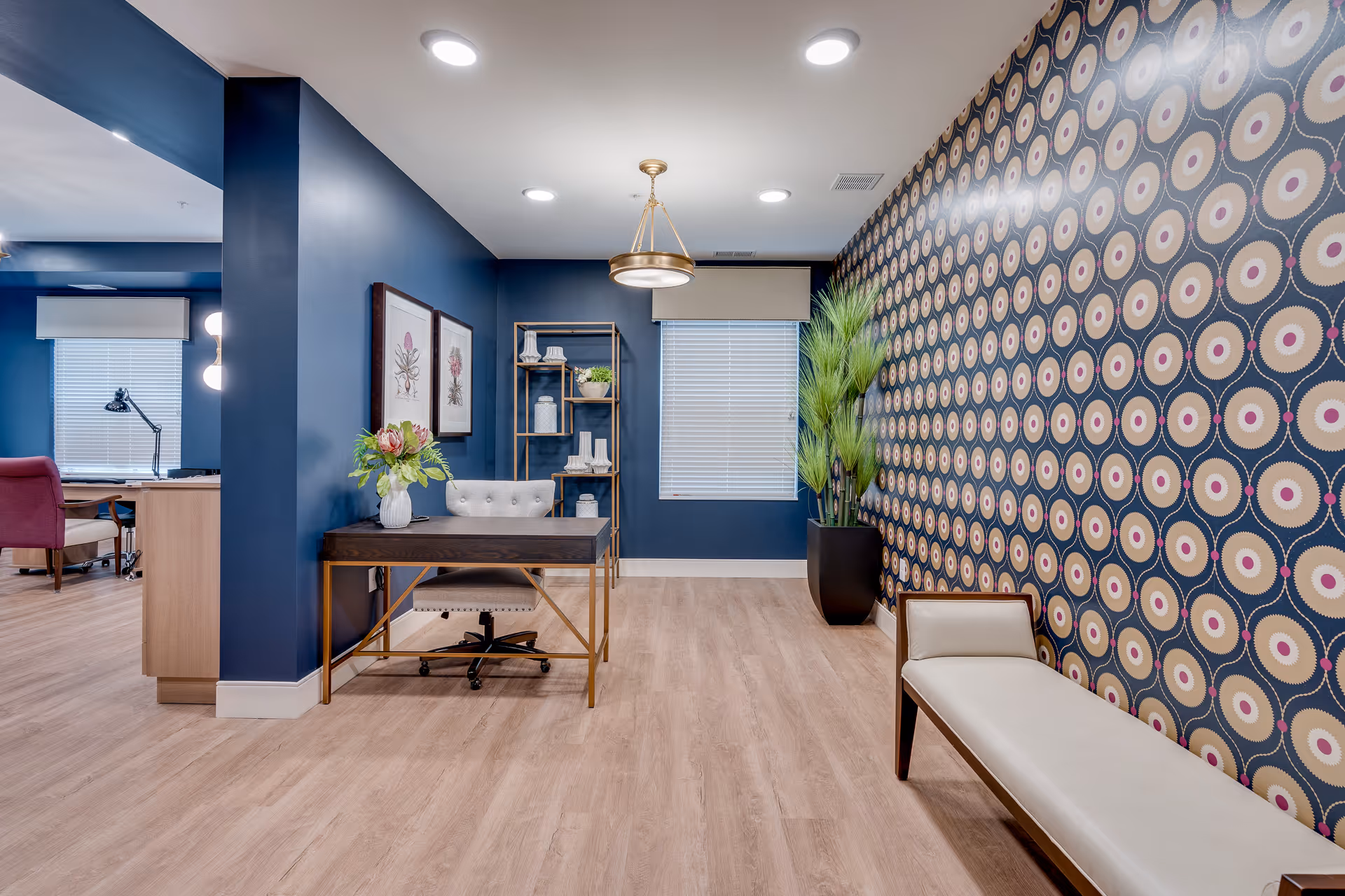 A modern interior space with blue walls and light wood flooring. On the right, there is a patterned wallpaper with circular designs and a white cushioned bench with wooden legs. In the center, a dark wooden desk with a white cushioned chair and a vase with flowers on top. Behind the desk, two framed botanical prints hang on the wall, and a tall shelving unit with decorative items is placed near a window with closed blinds. A large potted plant is positioned near the window. The ceiling has recessed lighting and a hanging light fixture.