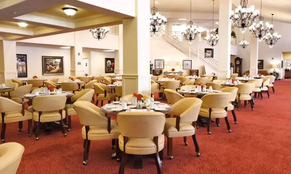 A spacious dining room with multiple round tables surrounded by beige upholstered chairs. Each table is set with white plates, cups, and silverware, and decorated with small flower arrangements. The room has red carpet flooring, white walls, and several chandeliers hanging from the ceiling. There are framed paintings on the walls and a staircase visible in the background.