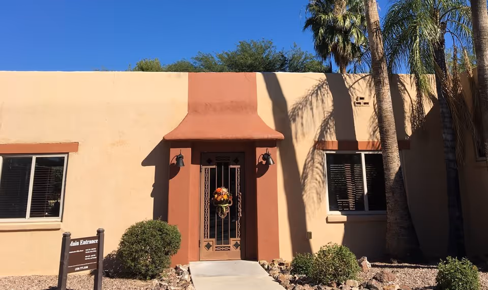Single-story stucco building front showing a central ornate metal door with a wreath, flanked by windows, shrubs, and palm trees.