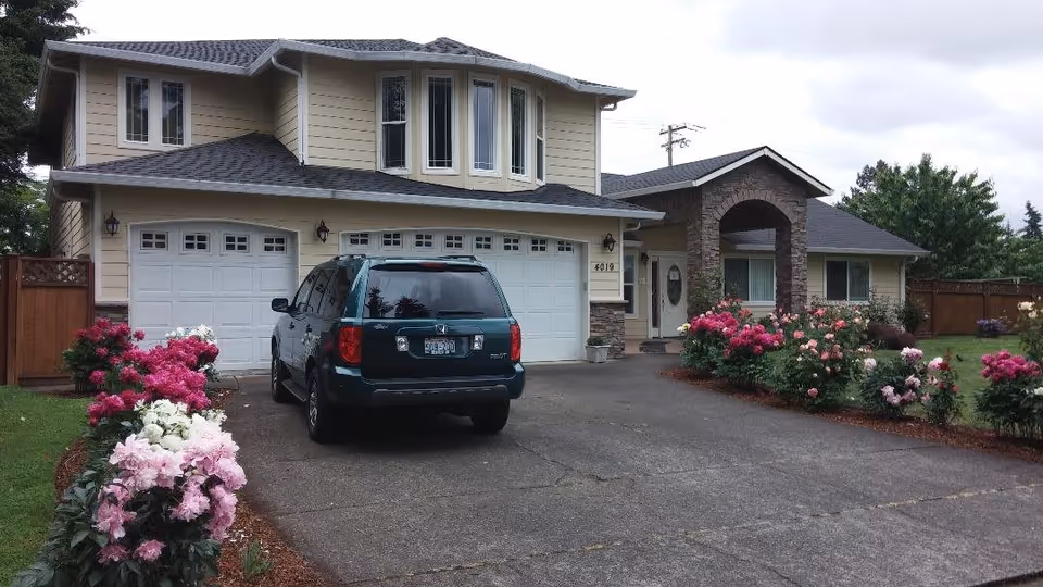 Front exterior view of a two-story house with beige siding and stone accents, a three-car garage, a driveway with a dark green SUV parked, and landscaped flower beds with pink and white flowers on either side of the driveway.