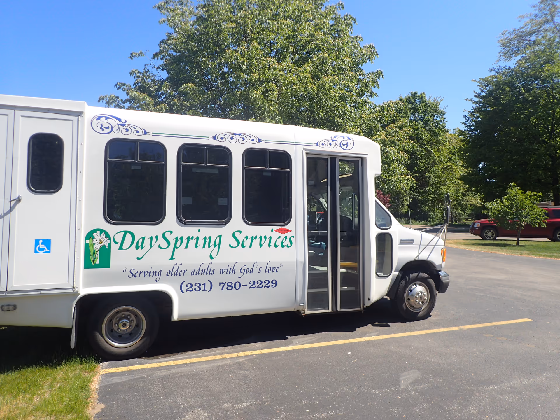 A white shuttle bus parked in a parking lot with trees and a red vehicle in the background. The bus has the text 'DaySpring Services' along with a phone number and the slogan 'Serving older adults with God's love' on its side. There is also a handicap accessibility symbol near the rear door.