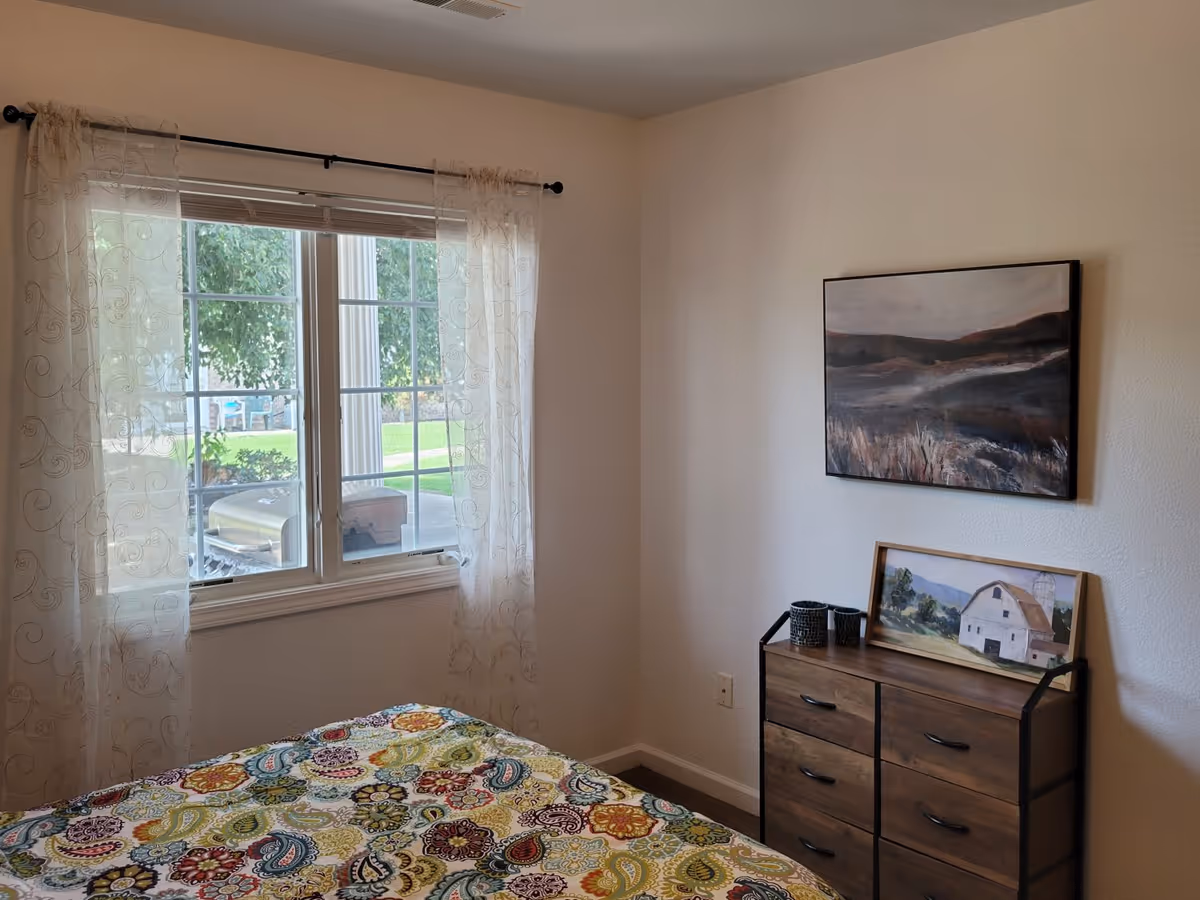 A bedroom with a window covered by sheer curtains, a bed with a colorful patterned bedspread, and a wooden dresser with two paintings and two small decorative containers on top.
