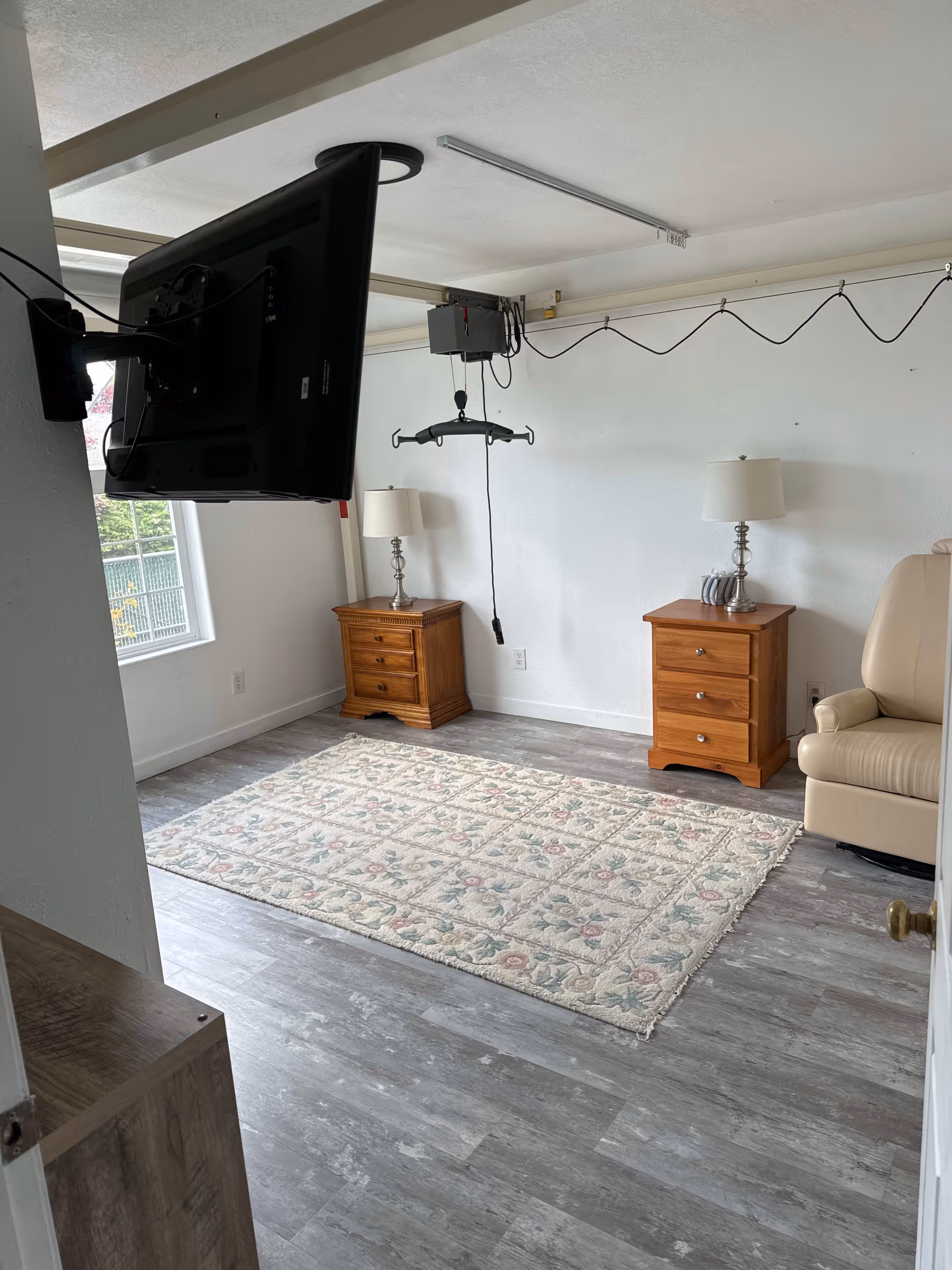 A small living room with two wooden nightstands each holding a lamp, a beige recliner chair, a floral patterned area rug on gray wood flooring, a wall-mounted TV, and a ceiling lift system with a sling bar. A window with a view of greenery is visible on the left side.