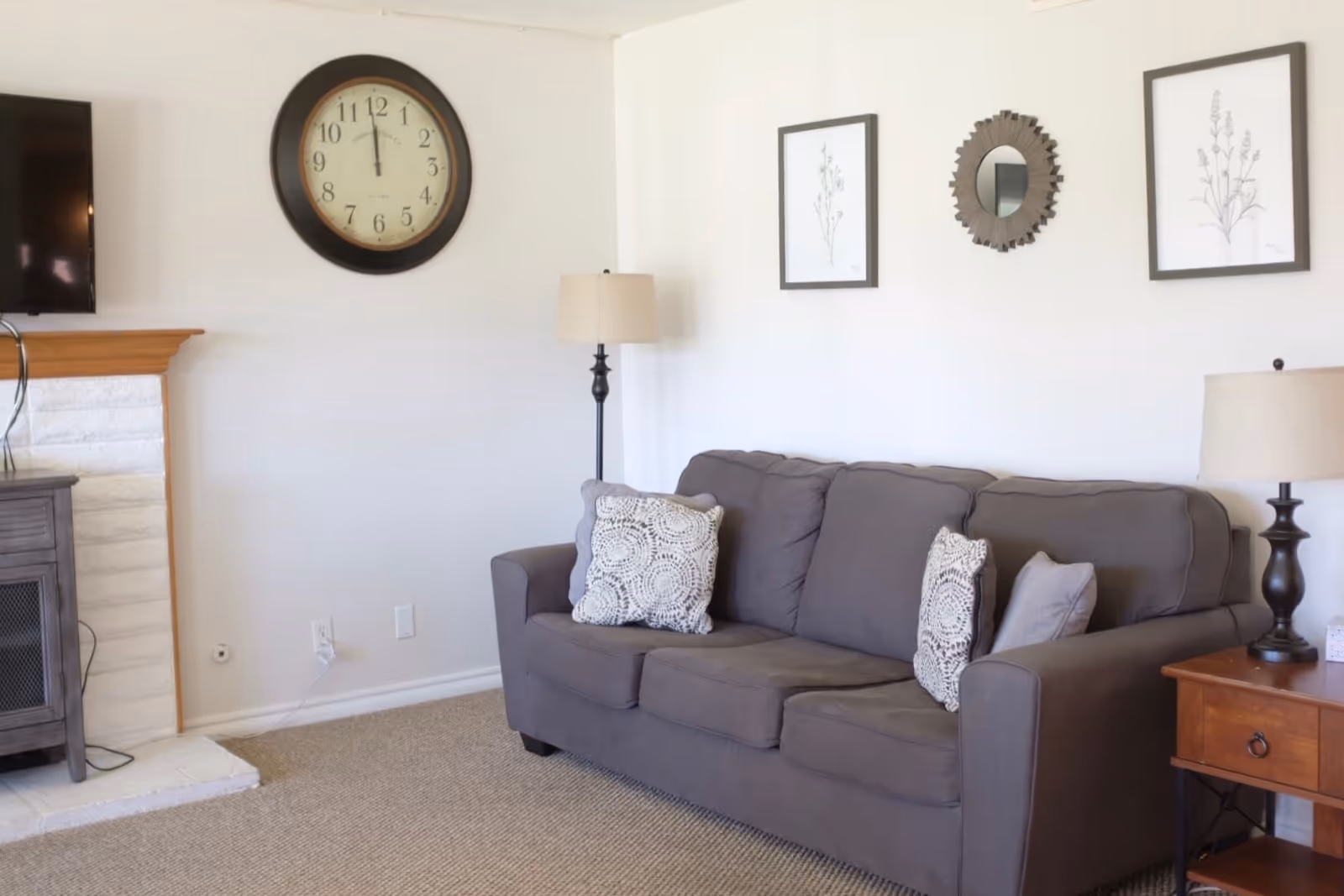 A cozy living room with a gray three-seater sofa adorned with patterned cushions. On the wall above the sofa are two framed botanical prints and a decorative round mirror. To the right of the sofa is a wooden side table with a lamp. On the left side of the image, there is a fireplace with a wooden mantel and a large round clock hanging above it. The room has light-colored walls and carpeted flooring.