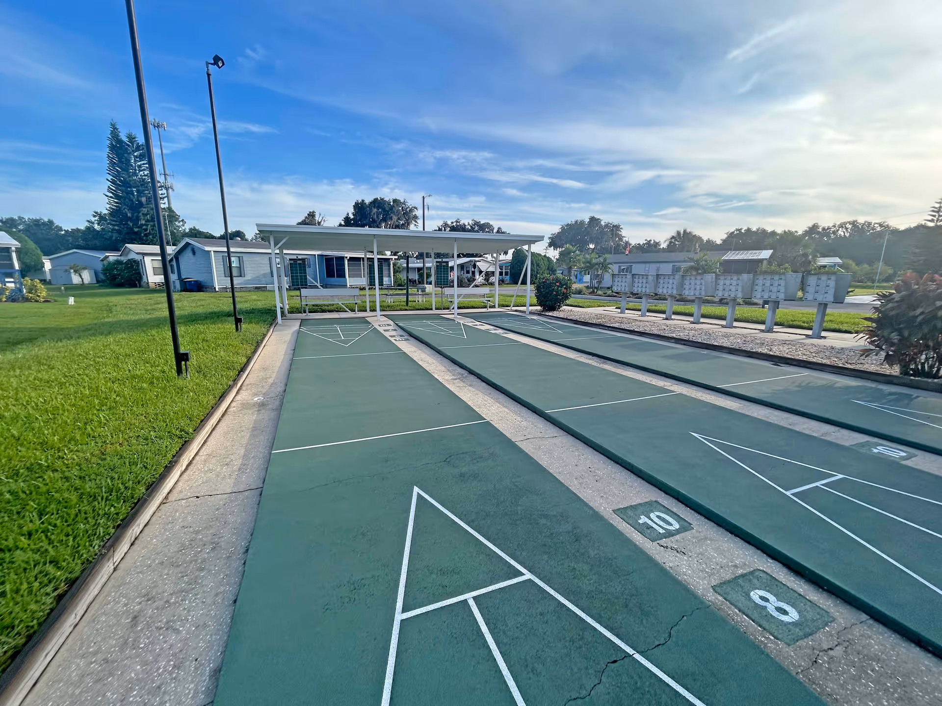 Shuffleboard courts at Oak Hammock Retirement Community with a covered pavilion, mailboxes, and nearby homes under a blue sky.