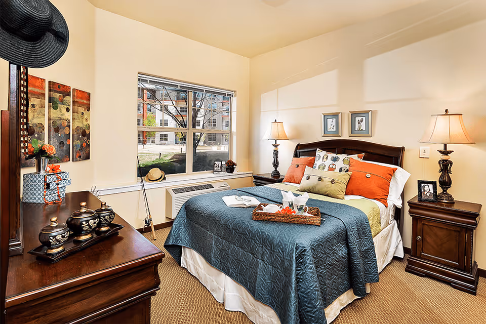 A cozy bedroom in a senior living facility with a neatly made bed featuring a blue quilt and multiple colorful pillows. The room has two wooden nightstands with lamps, framed pictures on the wall, a window showing an outdoor view, and a wooden dresser with decorative items including a hat and small containers.