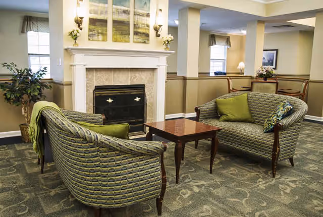 A cozy senior living facility common area with two patterned sofas facing each other around a wooden coffee table. There is a fireplace with a white mantel and decorative artwork above it. The room has beige walls, carpeted floor, and windows with curtains allowing natural light in. A small table with chairs and a lamp is visible in the background.