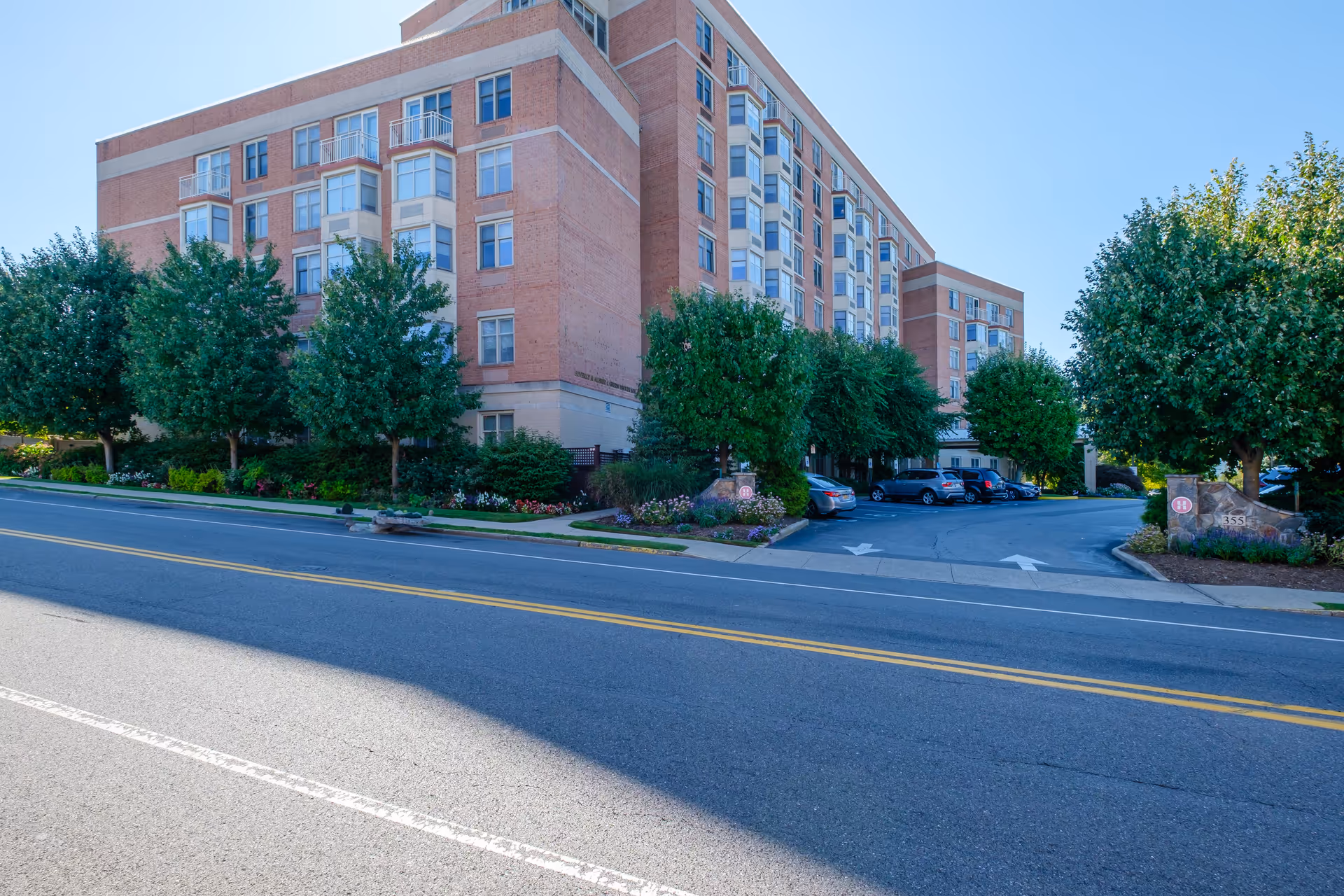 Exterior view of a multi-story brick building with numerous windows and balconies, surrounded by green trees and landscaped flower beds. There is a parking lot with several cars to the right of the building, and a clear blue sky overhead.