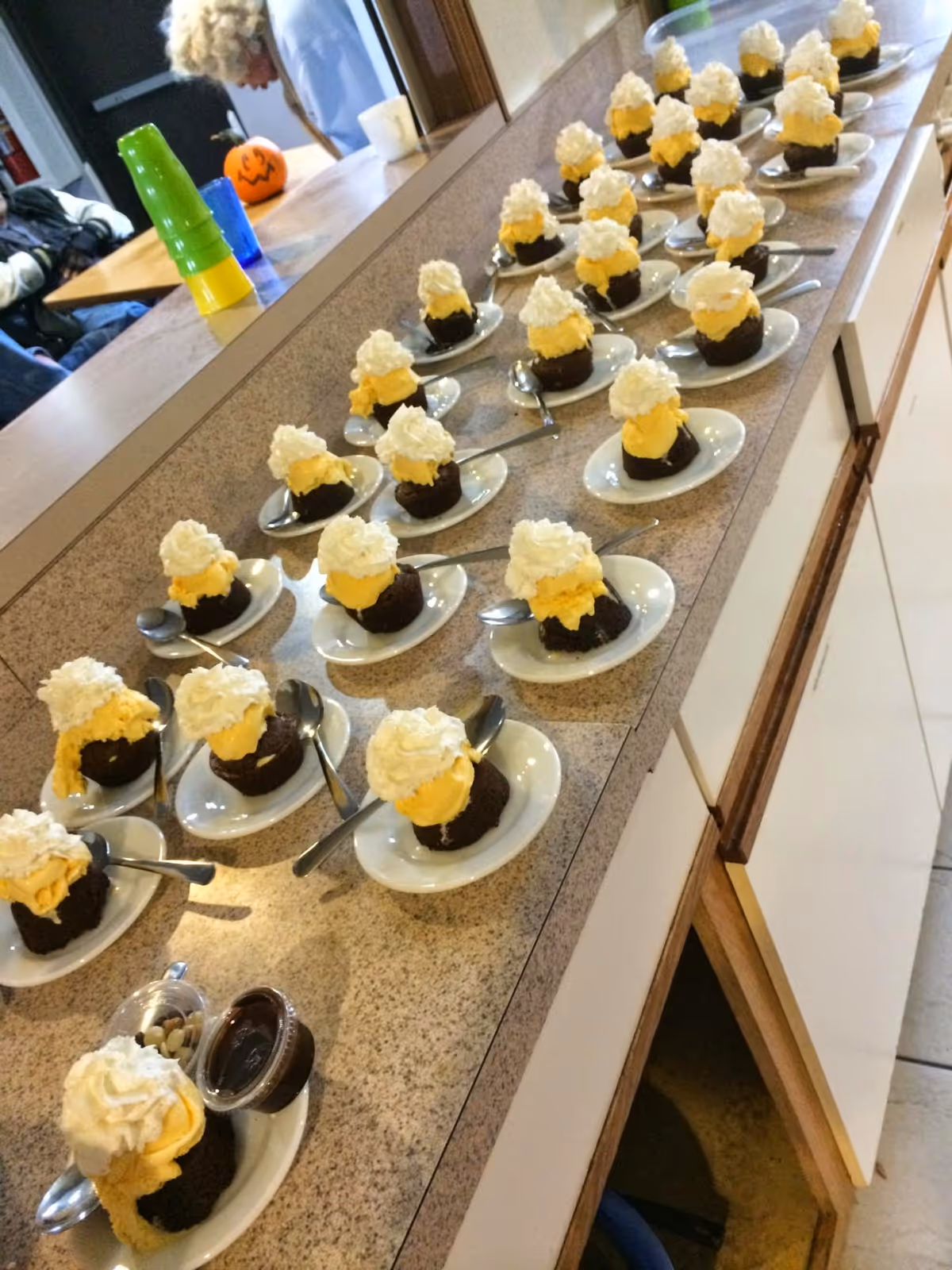 A countertop lined with multiple small plates, each holding a dessert consisting of a chocolate base topped with yellow ice cream and whipped cream, with a spoon placed on each plate. In the background, a person with curly white hair is seen near a table with a small pumpkin decoration and colorful plastic cups stacked together.