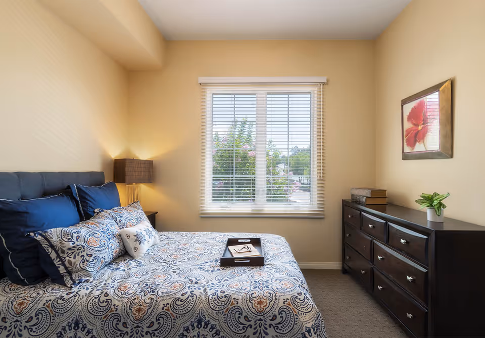 A bedroom with a bed covered in a blue and white patterned bedspread and multiple pillows. There is a wooden nightstand with a lamp on the left side of the bed. A window with white blinds is centered on the wall opposite the bed, showing greenery outside. On the right side of the room, there is a dark wooden dresser with books, a small plant, and a framed flower artwork hanging above it.