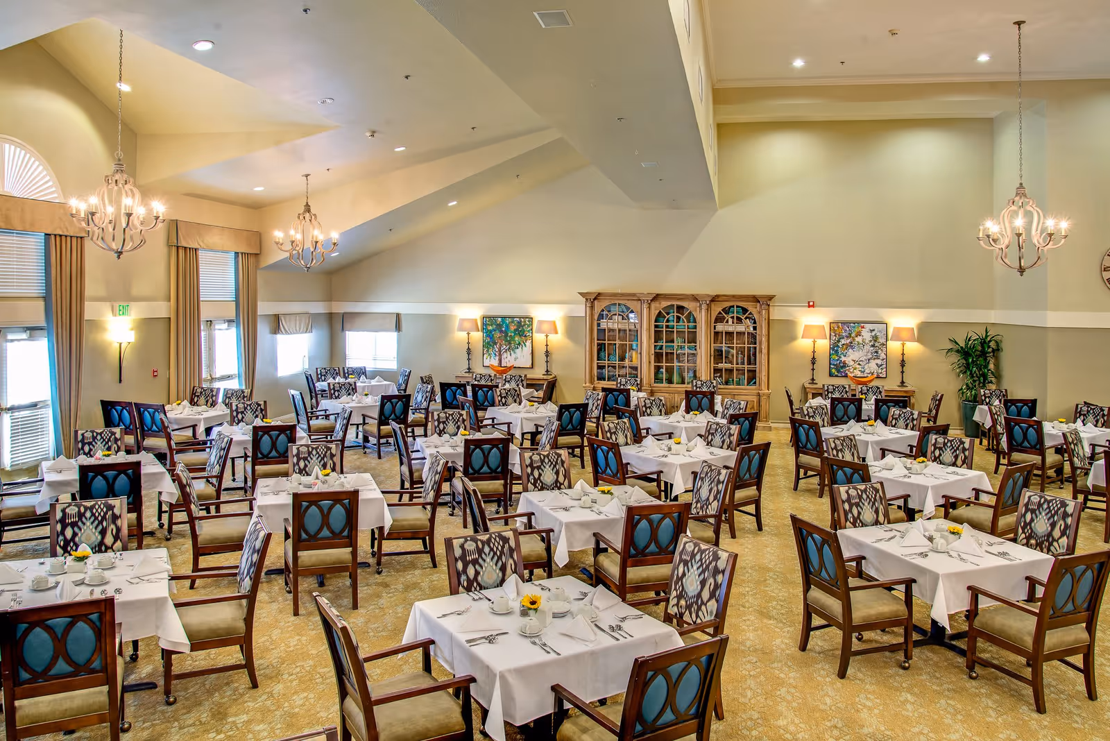 A spacious dining room with multiple tables covered in white tablecloths, each set with cups, plates, and silverware. The room features patterned chairs, chandeliers hanging from a high ceiling, large windows with curtains, and decorative lamps and artwork on the walls.