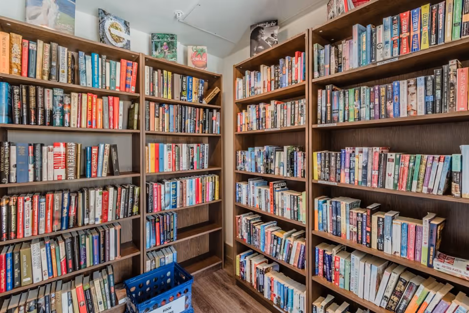 Interior view of a small library or reading room with multiple wooden bookshelves filled with books. There is a blue plastic crate on the floor labeled 'BOOK RETURNS'.