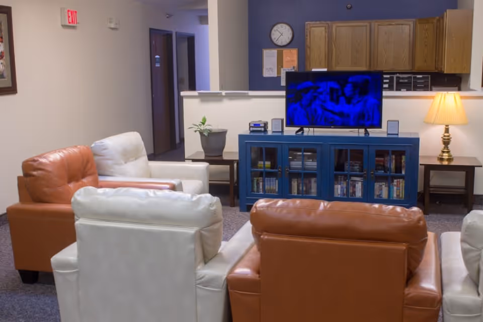 A cozy living room area with four leather armchairs, two white and two brown, arranged facing a blue TV stand with a flat-screen television on top. The TV stand has glass doors revealing books and DVDs inside. Behind the TV stand is a half wall with a kitchen area visible, featuring wooden cabinets and a lamp on a side table. A clock and bulletin board are mounted on the blue wall above the TV stand.