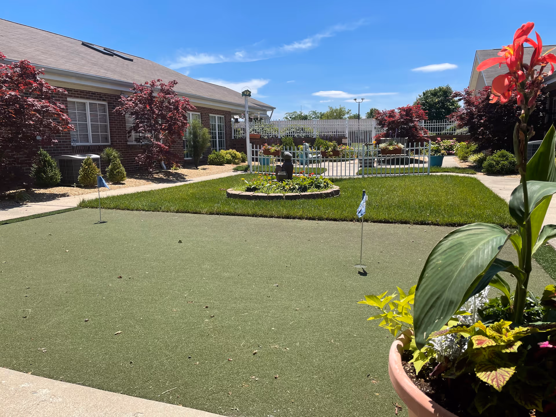 Sunny courtyard with a small putting green, flower beds, potted plants, and a brick building in the background.