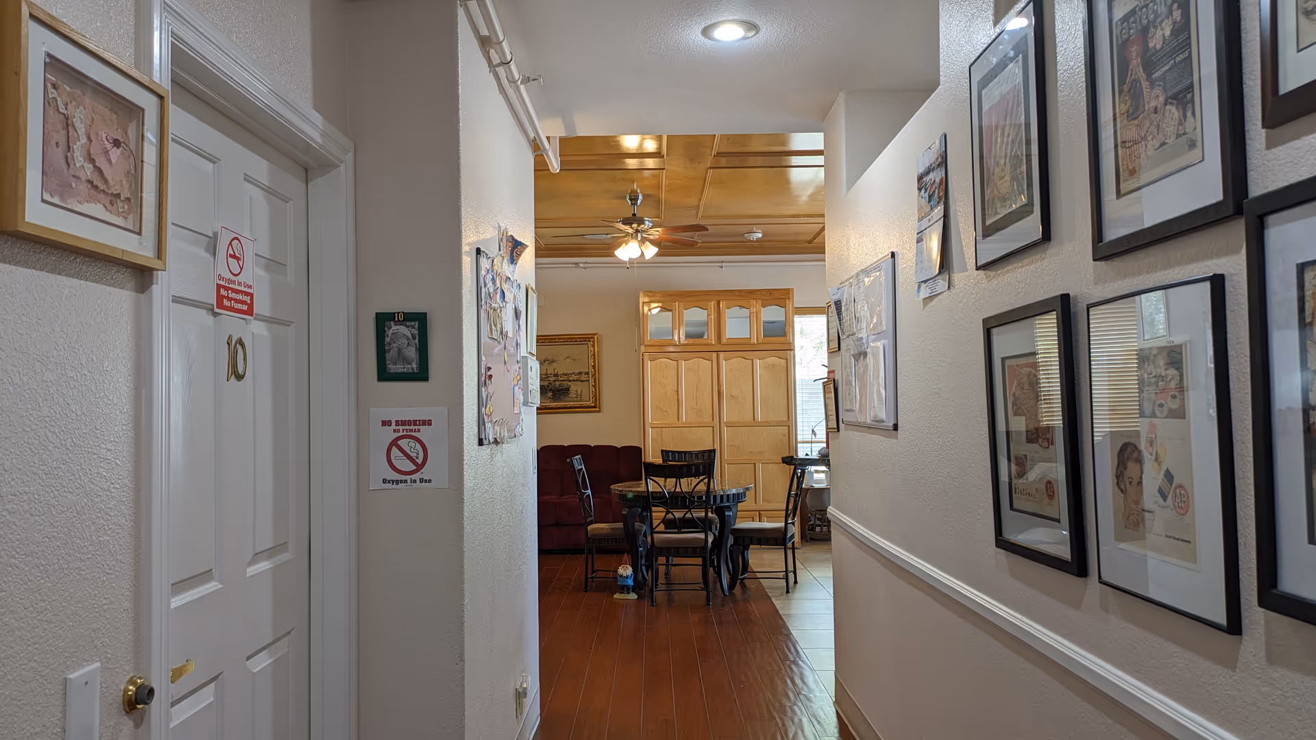 Interior hallway of a senior living facility with a white door labeled number 10 on the left, walls decorated with framed pictures and notices, leading to a room with a round table and chairs, a ceiling fan, and a wooden cabinet. The hallway has wooden flooring and a white ceiling with a recessed light.