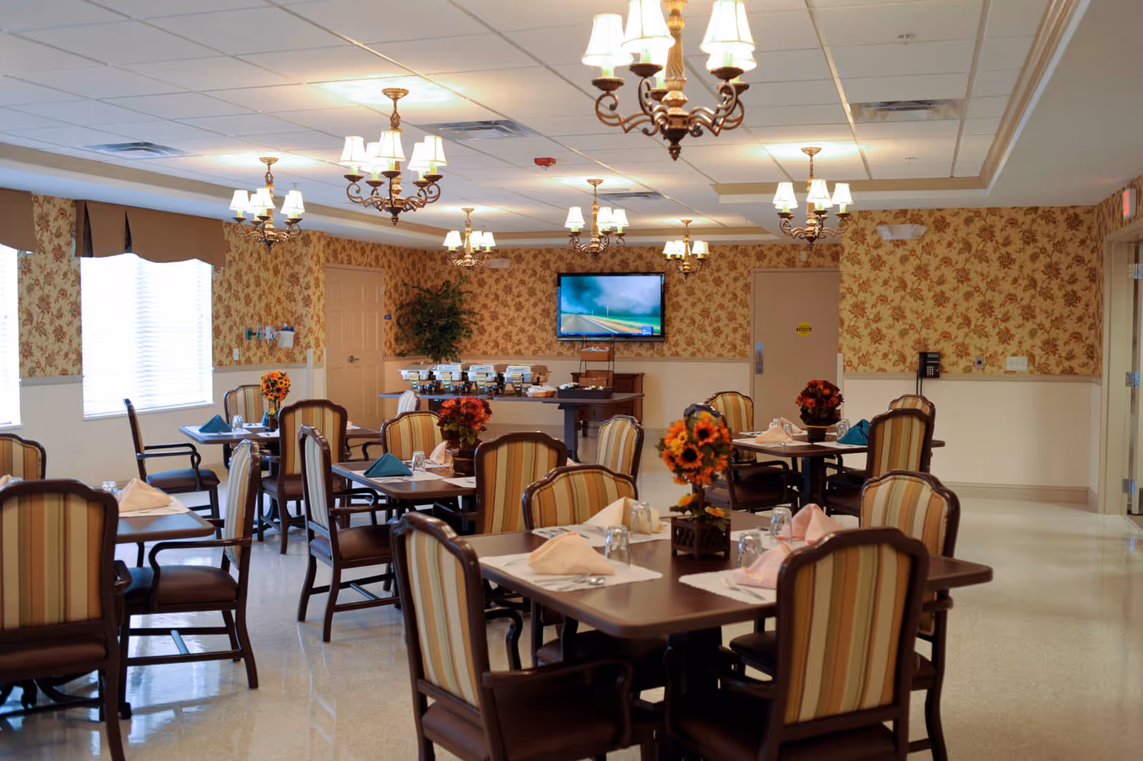 Bright dining room with multiple tables and striped chairs, chandeliers, floral centerpieces, and a TV on the far wall.