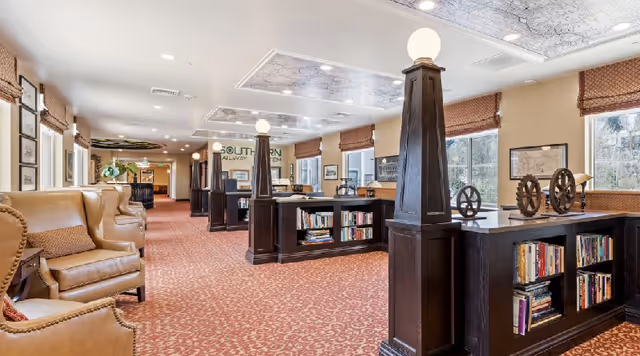 Bright, carpeted common lounge with leather armchairs, dark wood bookcases, decorative lighted columns and windows.