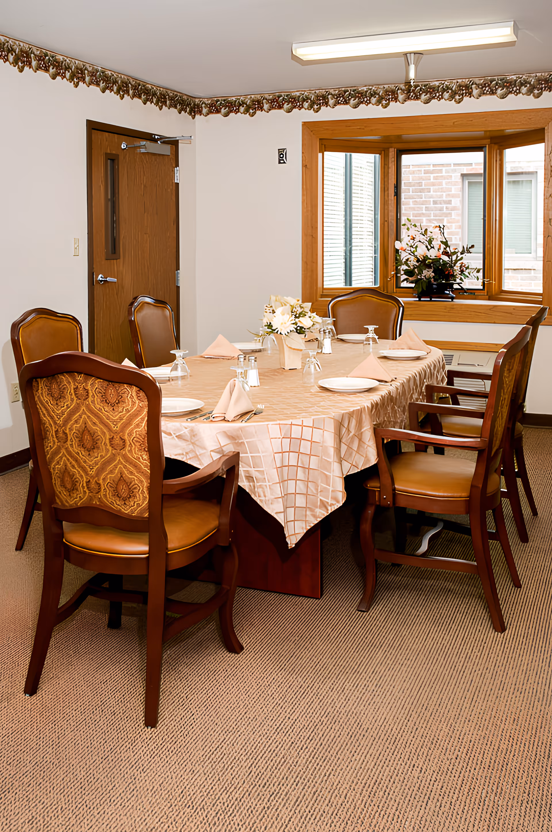 A dining room with a rectangular table covered with a beige tablecloth. The table is set with plates, folded napkins, upside-down glasses, and silverware. There are six wooden chairs with cushioned seats around the table. A window with wooden trim and a flower arrangement is visible in the background. The room has beige carpet, white walls, and a wooden door.