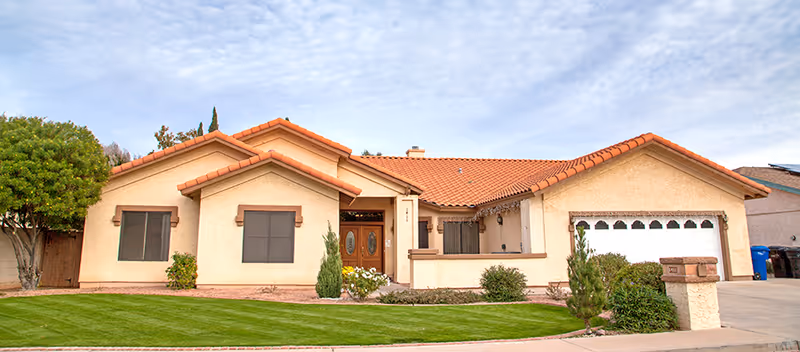 Single-story beige house with red tile roof and manicured lawn in front.