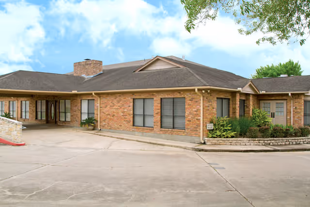 Single-story brick senior living building with a covered entry, wrap-around driveway, and landscaped planter under a blue sky.