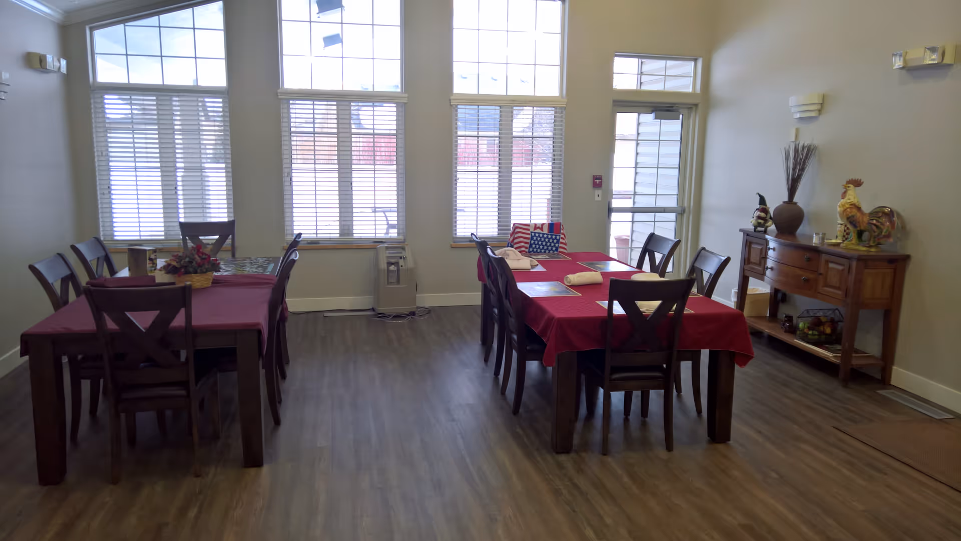 A dining room with two wooden tables covered with red tablecloths, each surrounded by wooden chairs. The room has large windows with blinds letting in natural light. There is a wooden sideboard against the right wall decorated with a rooster figurine, a vase, and other small items. The floor is wood, and the walls are light-colored.