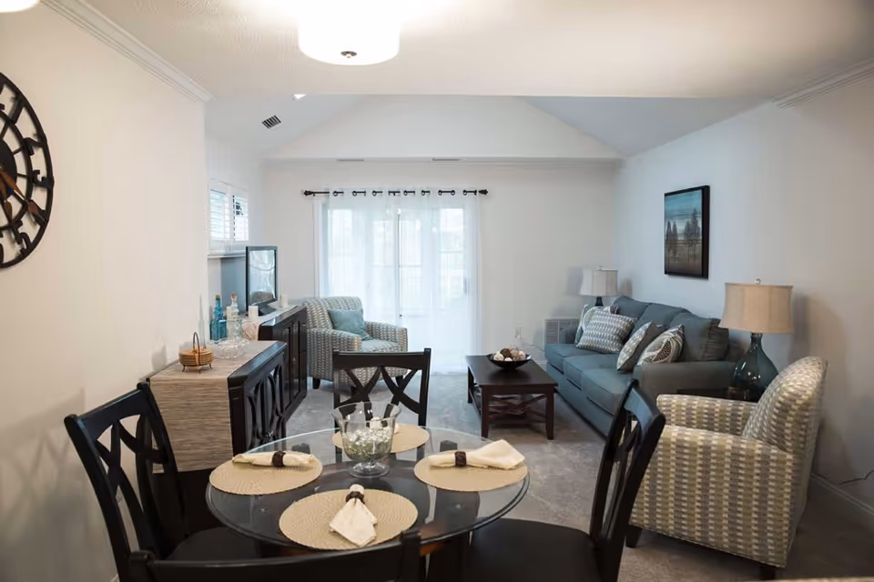A bright and cozy living and dining area in a retirement community. The dining area features a round glass table set with four place settings, each with a beige placemat and a folded napkin. The living area includes a blue sofa with several patterned cushions, two armchairs with a geometric pattern, a dark wooden coffee table with decorative items, and a sideboard with bottles and glassware. Large windows with sheer white curtains allow natural light to fill the room. The walls are painted white and decorated with a large round clock and a framed landscape painting.
