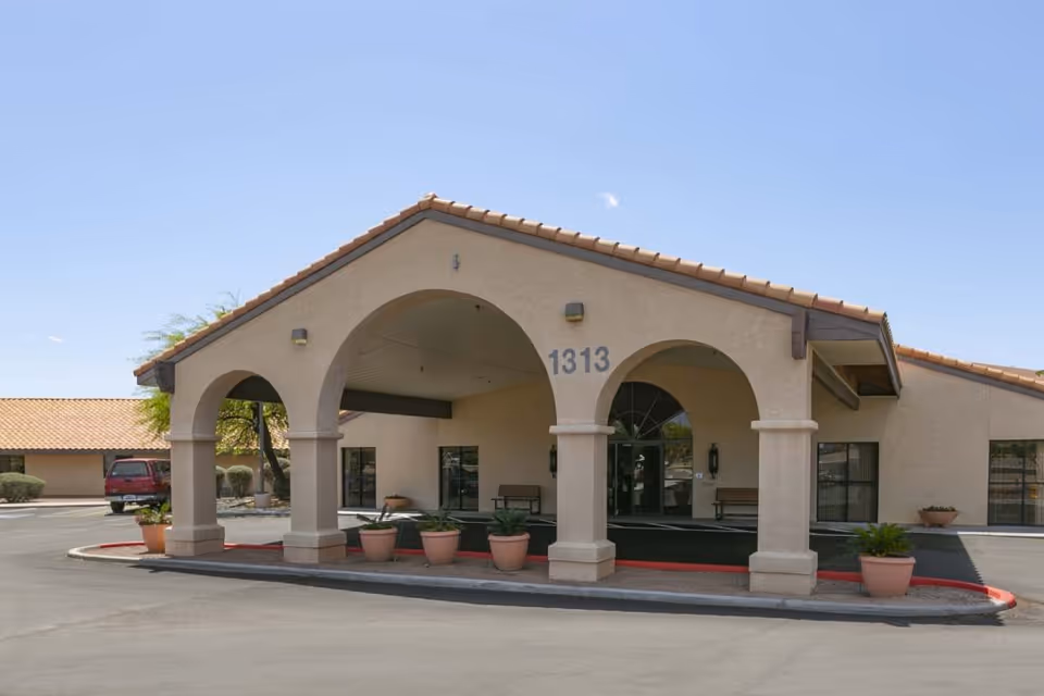 Exterior view of Mountain View Care Center showing the entrance with three large arches, beige stucco walls, and a tiled roof. Several potted plants are placed near the entrance, and the building number 1313 is visible above the middle arch. A red curb lines the driveway in front of the entrance under a clear blue sky.